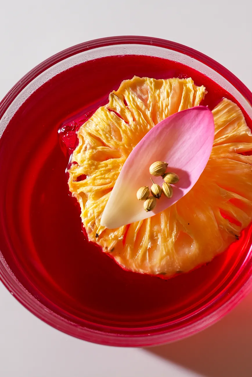A dried pineapple slice topped with a lotus petal in a red drink